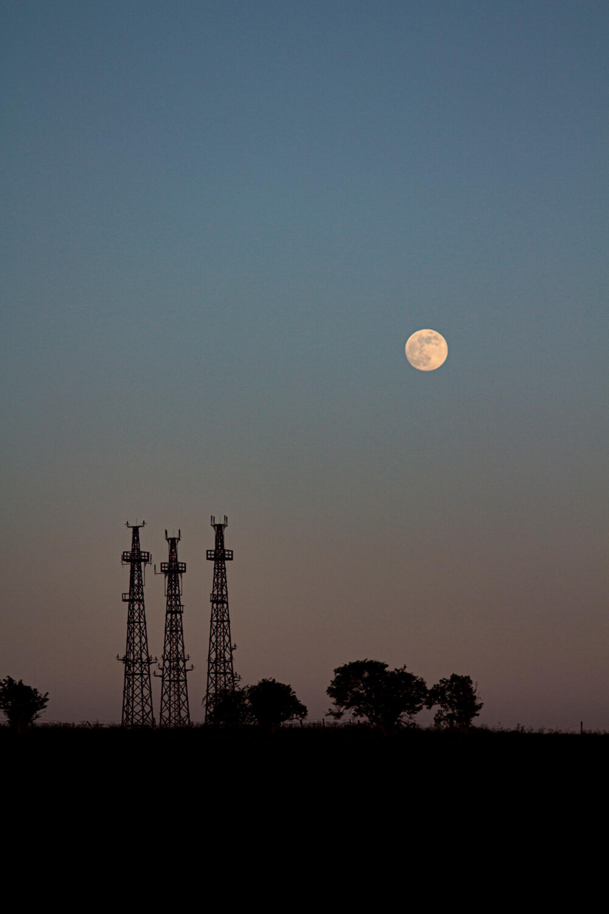 Landscape shot at dusk where the sky is an orange to blue gradient with the moon visible. On the horizon are some small trees and electrical towers